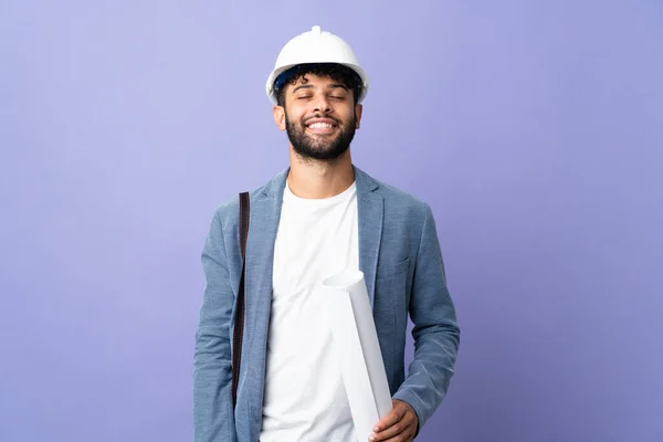 Young architect Moroccan man with helmet and holding blueprints over isolated background laughing