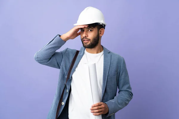 Young architect Moroccan man with helmet and holding blueprints over isolated background doing surprise gesture while looking to the side