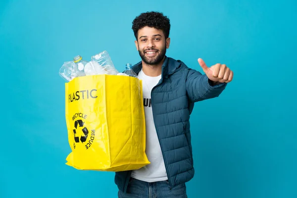 Young Moroccan man holding a bag full of plastic bottles to recycle over isolated background giving a thumbs up gesture