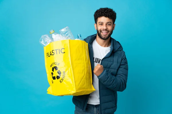 Young Moroccan man holding a bag full of plastic bottles to recycle over isolated background celebrating a victory