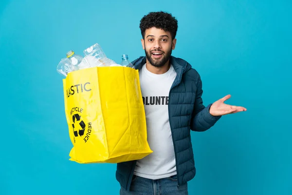 Young Moroccan man holding a bag full of plastic bottles to recycle over isolated background with shocked facial expression