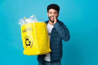 Young Moroccan man holding a bag full of plastic bottles to recycle over isolated background shouting with mouth wide open