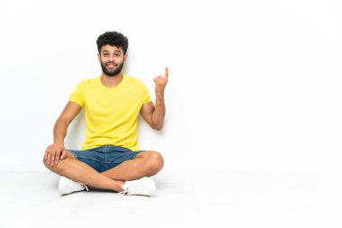 Young Moroccan handsome man sitting on the floor over isolated background pointing with the index finger a great idea