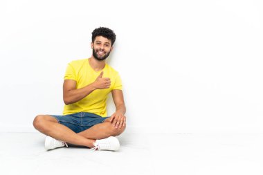 Genç Faslı yakışıklı adam, izole edilmiş arka planda yerde oturuyor ve baş parmak işareti yapıyor.