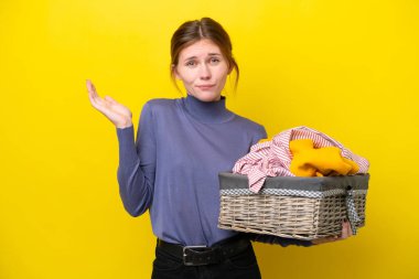 Young English woman holding a clothes basket isolated on yellow background having doubts while raising hands