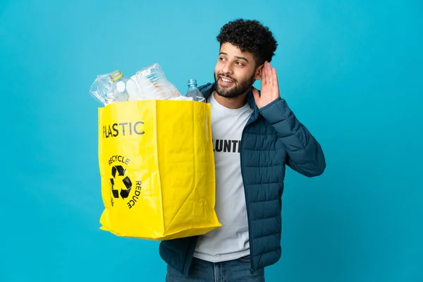 Young Moroccan man holding a bag full of plastic bottles to recycle over isolated background listening to something by putting hand on the ear