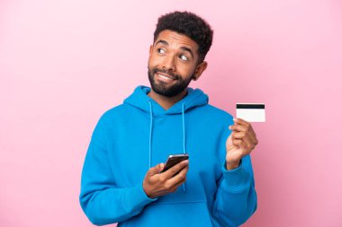 Young Brazilian man isolated on pink background buying with the mobile with a credit card while thinking
