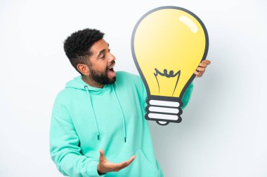 Beyaz arka planda izole edilmiş genç Brezilyalı bir adam elinde şaşırmış bir ifadeyle bir ampul simgesi tutuyor.