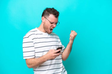 Genç Brezilyalı adam mavi arka planda izole edilmiş ve telefon zafer pozisyonunda.