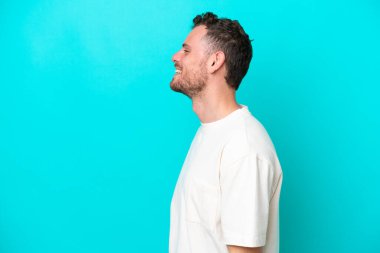 Genç Brezilyalı adam mavi arka planda izole edilmiş. Yan pozisyonda gülüyor.