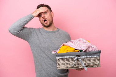 Genç Brezilyalı bir adam pembe arka planda izole edilmiş bir çamaşır sepeti tutuyor. Yan tarafa bakarken sürpriz jest yapıyor.