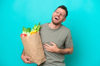 Elinde alışveriş çantası tutan Brezilyalı genç adam mavi arka planda izole edilmiş, gülümsüyor.