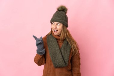 Kış şapkalı orta yaşlı kadın pembe arka planda izole edilmiş. Çözümün ne olduğunu anlamak için parmağını kaldırıyor.