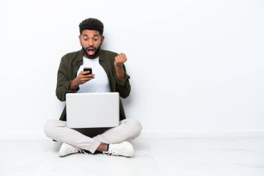 Yerde dizüstü bilgisayarlı genç Brezilyalı bir adam oturuyor. Beyazlara izole edilmiş, şaşırmış ve mesaj yolluyor.