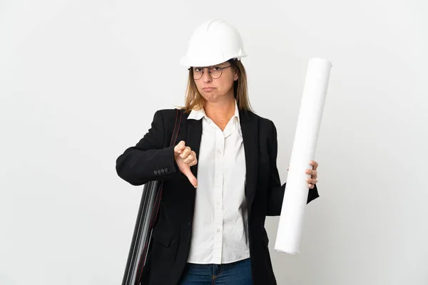Middle age architect woman with helmet and holding blueprints over isolated background showing thumb down with negative expression