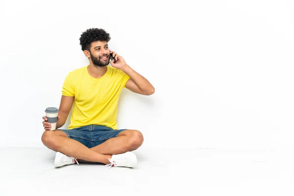 Young Moroccan handsome man sitting on the floor over isolated background holding coffee to take away and a mobile