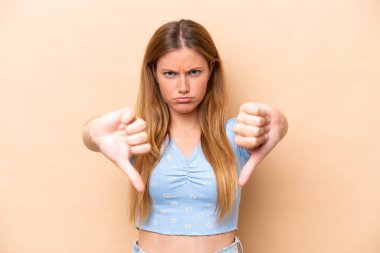 Beyazlı genç bir kadın bej arka planda izole edilmiş, baş parmağını iki eliyle aşağı gösteriyor.