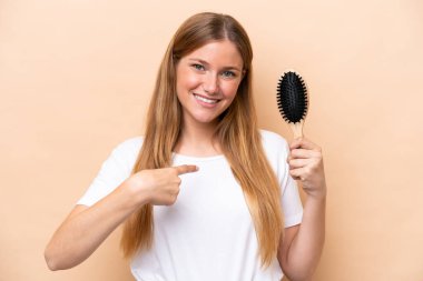 Young pretty blonde woman with hair comb isolated on beige background with surprise facial expression