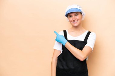 Balıkçı beyaz kadın önlük giyiyor ve bej arka planda izole edilmiş çiğ balık tutuyor. Ürünü sunmak için yan tarafı işaret ediyor.