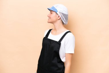 Balıkçı beyaz kadın önlük takıyor ve bej arka planda izole edilmiş çiğ bir balığı tutuyor. Yan pozisyonda gülüyor.