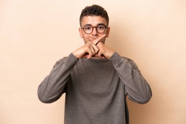 Bej arka planda izole edilmiş genç Brezilyalı bir adam sessizlik işareti gösteriyor.