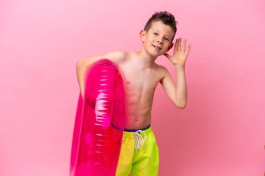 Küçük beyaz bir çocuk pembe arka planda izole edilmiş şişme bir çörek tutuyor. Kulağına elini sürerek bir şeyler dinliyor.