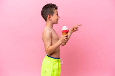 Pembe arka planda izole edilmiş dondurma yiyen beyaz bir çocuk bir ürün sunmak için yan tarafı işaret ediyor.