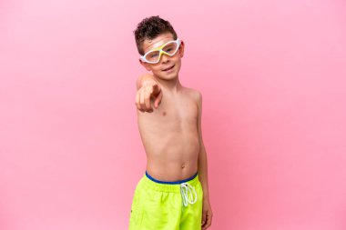 Pembe arka planda izole edilmiş dalış gözlüğü takan beyaz çocuk mutlu bir ifadeyle önü işaret ediyor.