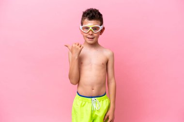 Pembe arka planda izole edilmiş dalış gözlüğü takan beyaz bir çocuk bir ürün sunmak için yan tarafı işaret ediyor.