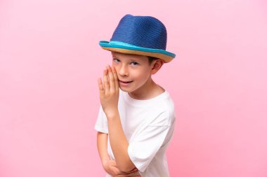 Pembe arka planda izole edilmiş şapka takan küçük beyaz çocuk bir şeyler fısıldıyor.