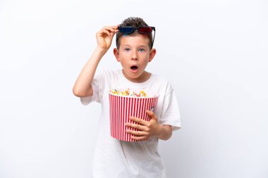 Beyaz arka planda izole edilmiş küçük beyaz çocuk 3D gözlüklü ve elinde koca bir kova patlamış mısırla şaşırdı.