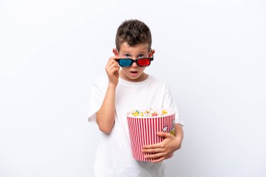 Beyaz arka planda izole edilmiş küçük beyaz çocuk 3D gözlüklü ve elinde koca bir kova patlamış mısırla şaşırdı.