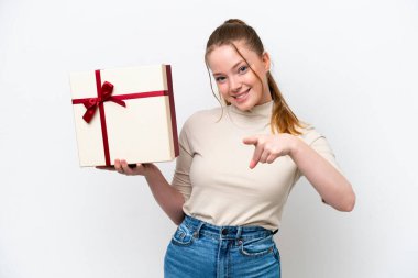 Beyaz arka planda izole edilmiş beyaz bir hediye tutan genç beyaz kadın kendine güvenen bir ifadeyle seni işaret ediyor.