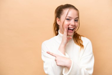 Beyaz tenli, bej arka planda izole edilmiş genç bir kadın bir ürünü sunmak ve bir şeyler fısıldamak için kenarı işaret ediyor.