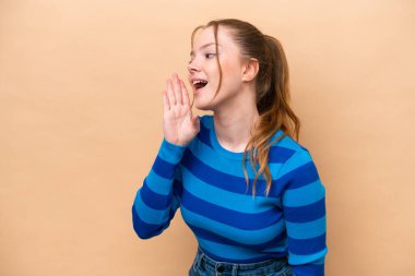 Beyaz tenli genç bir kadın bej arka planda izole edilmiş ve ağzı sonuna kadar açık bağırıyor.