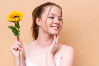 Beyaz tenli genç bir kız bej arka planda izole edilmiş, elinde bir ayçiçeği tutarken gülümsüyor. Portreyi kapat