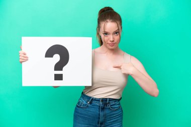 Young caucasian woman isolated on green background holding a placard with question mark symbol with surprised expression