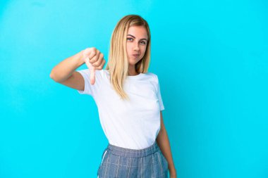 Sarışın Uruguaylı kız mavi arka planda izole edilmiş, baş parmağını negatif ifadeyle aşağı gösteriyor.