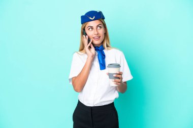 Uçak hostesi Uruguaylı kadın mavi arka planda izole edilmiş. Elinde kahve ve cep telefonu var.