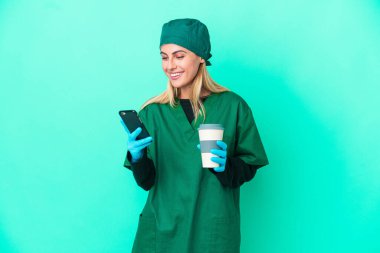 Yeşil üniformalı genç cerrah Uruguaylı kadın mavi arka planda izole edilmiş. Elinde kahve ve cep telefonu var.