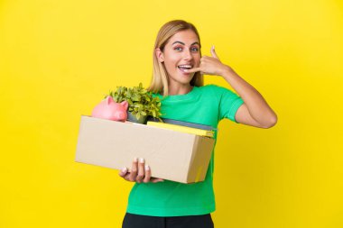 Genç Uruguaylı kız, sarı arka planda izole edilmiş eşyalarla dolu bir kutuyu alıp telefon hareketleriyle hareket ediyor. Bana geri dönüş işareti ver.