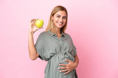 Genç Uruguaylı kadın mavi arka planda izole edilmiş hamile ve elinde bir elma var.