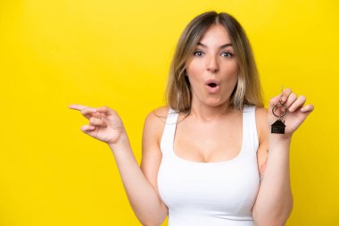 Sarı arka planda izole edilmiş ev anahtarlarını tutan genç Romen kadın şaşırmış ve işaret ediyor