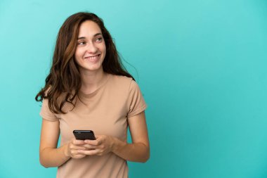 Kafkasyalı genç bir kadın mavi arka planda izole edilmiş ve cep telefonu kullanıyor.