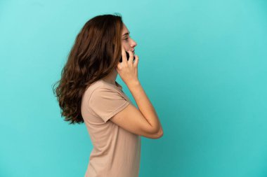 Mavi arka planda izole edilmiş genç beyaz bir kadın cep telefonuyla sohbet ediyor.