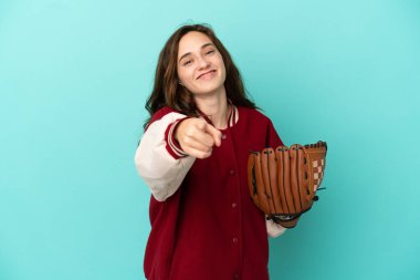 Beyzbol oynayan beyaz bir kadın mavi arka planda izole edilmiş, mutlu bir ifadeyle ön tarafı işaret ediyor.