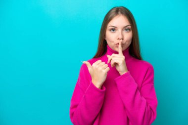 Genç Litvanyalı bir kadın mavi arka planda izole edilmiş, kenarı işaret ediyor ve sessizlik hareketi yapıyor.