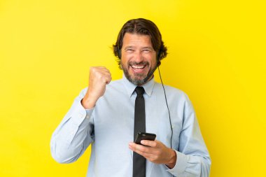 Tele pazarlamacı Hollandalı adam sarı arka planda izole edilmiş bir kulaklıkla çalışıyor ve telefon zafer pozisyonunda.