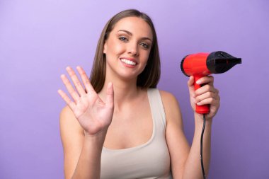 Young caucasian woman holding a hairdryer isolated on purple background saluting with hand with happy expression