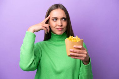 Young caucasian woman holding fried chips on purple background having doubts and thinking